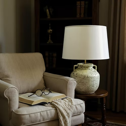 Cozy living room with a beige armchair, open book, glasses, and a lamp on a side table.