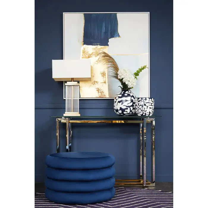 Decorative interior setting with a blue ottoman, glass table, and abstract art on a navy wall.