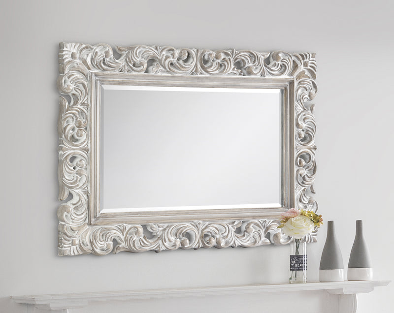 Decorative wall mirror with intricate white frame on a plain white wall.