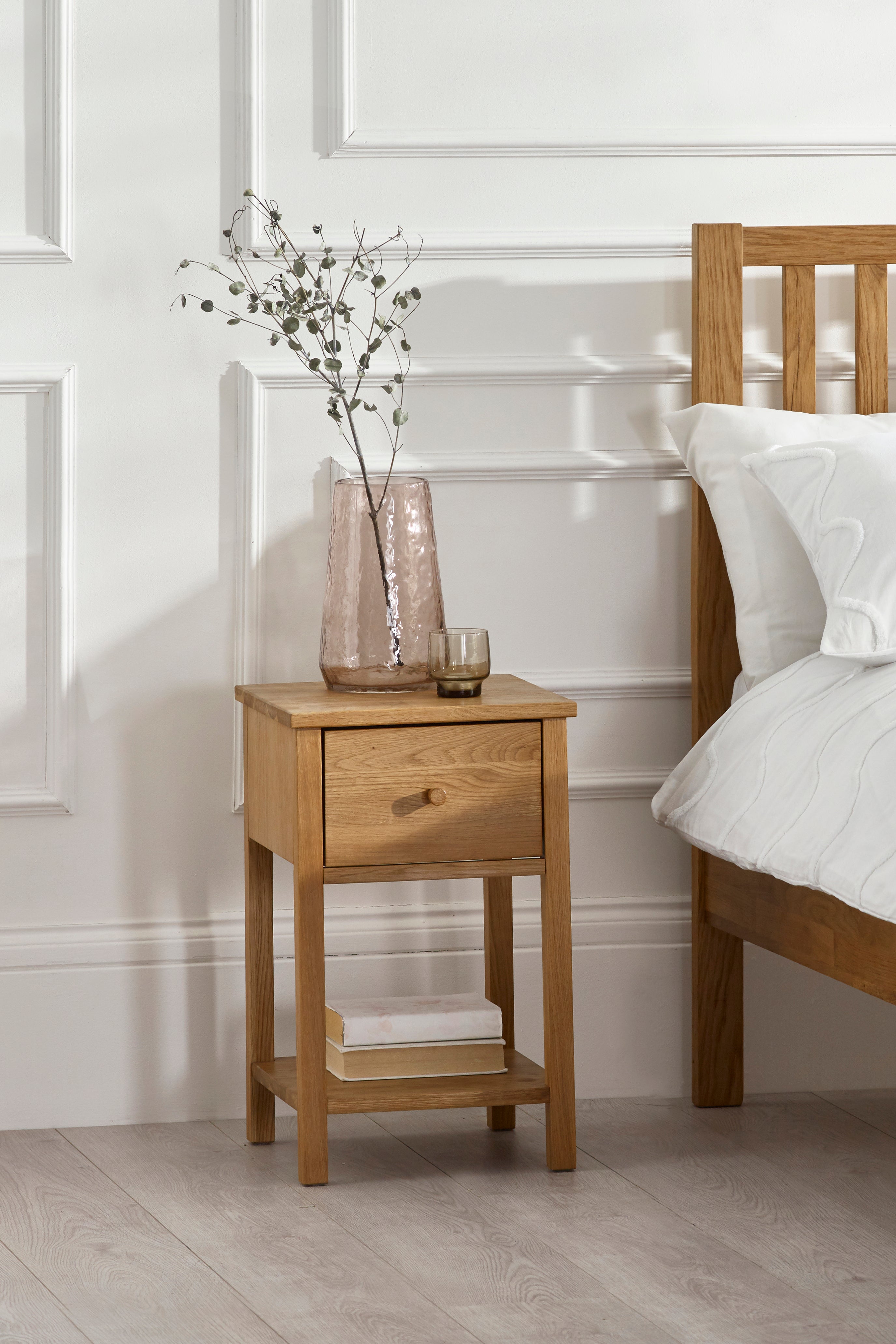 Wooden bedside table with a vase and books next to a bed in a bedroom.