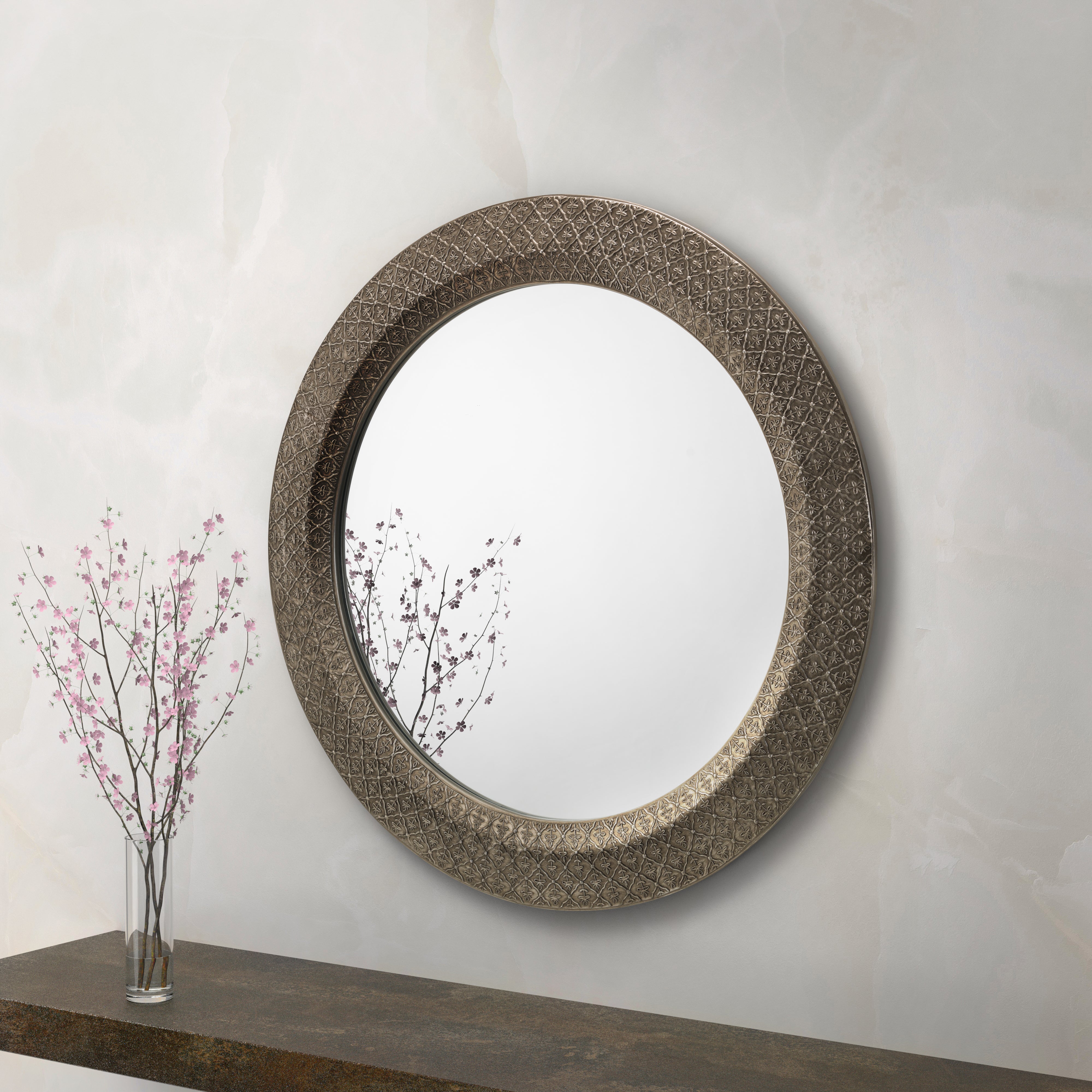 Round decorative mirror with intricate design on a white wall, next to a vase with pink branches.