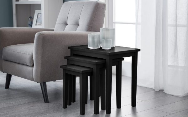 Set of black nesting tables in a living room setting with a gray armchair and white curtains.