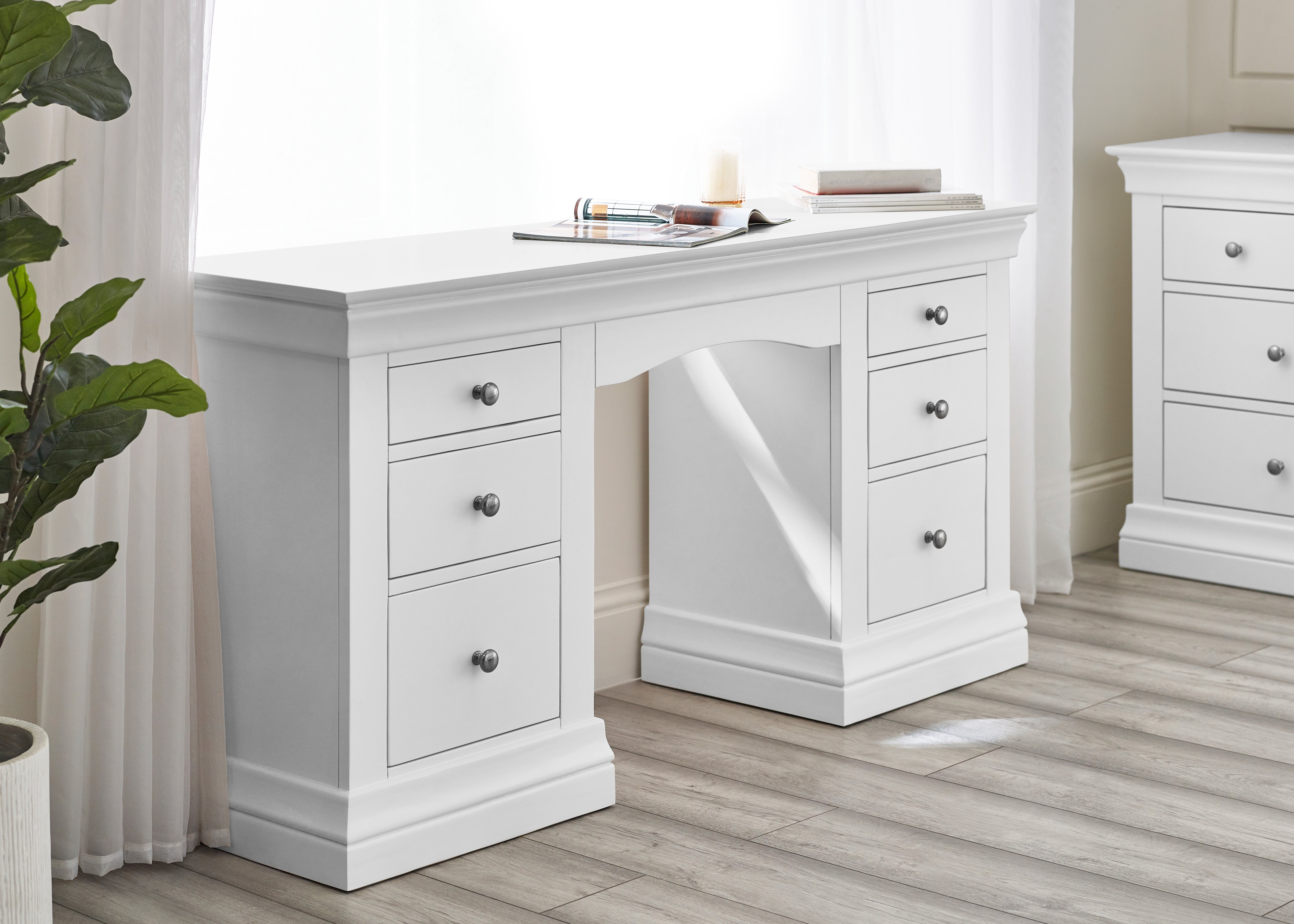 White desk with drawers in a room with curtains and a plant.
