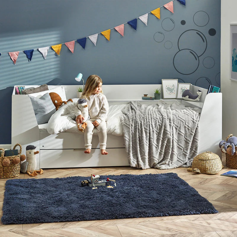 Child sitting on a white bed in a room with a blue wall, colorful flags, and toys.