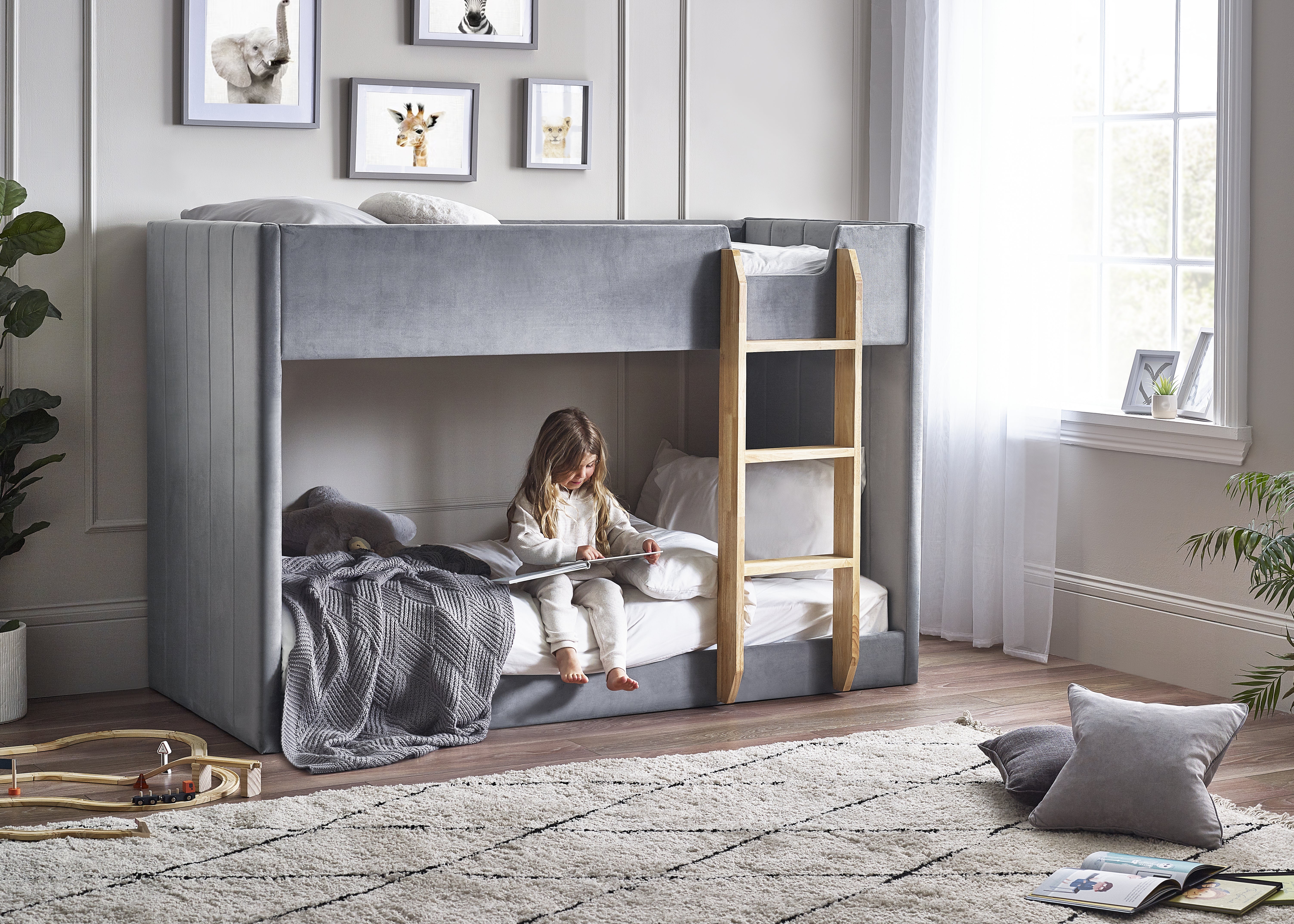 Child reading a book in a gray bunk bed with wooden steps in a bright room.
