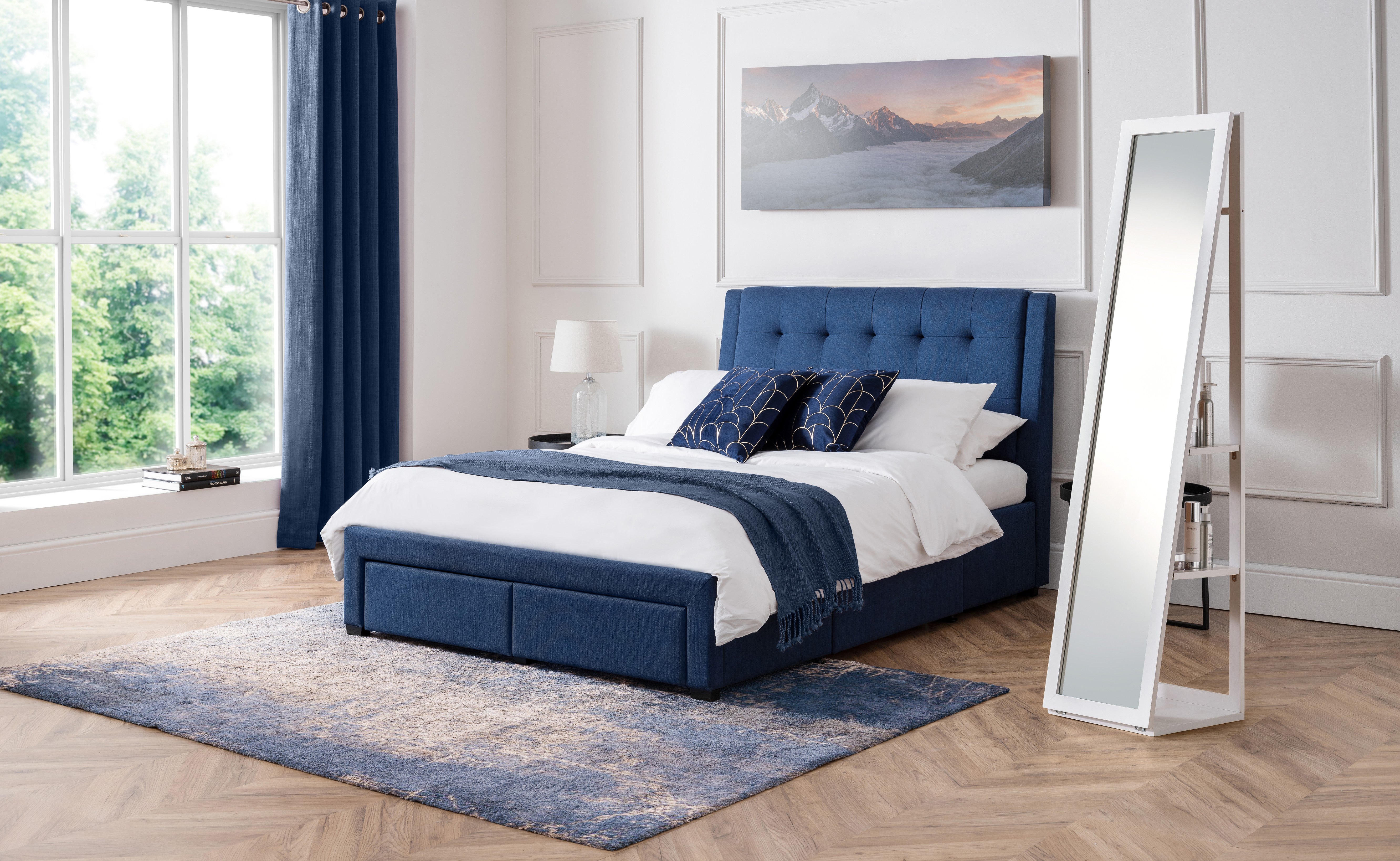 Blue upholstered bed in a bright bedroom with large windows and a standing mirror.