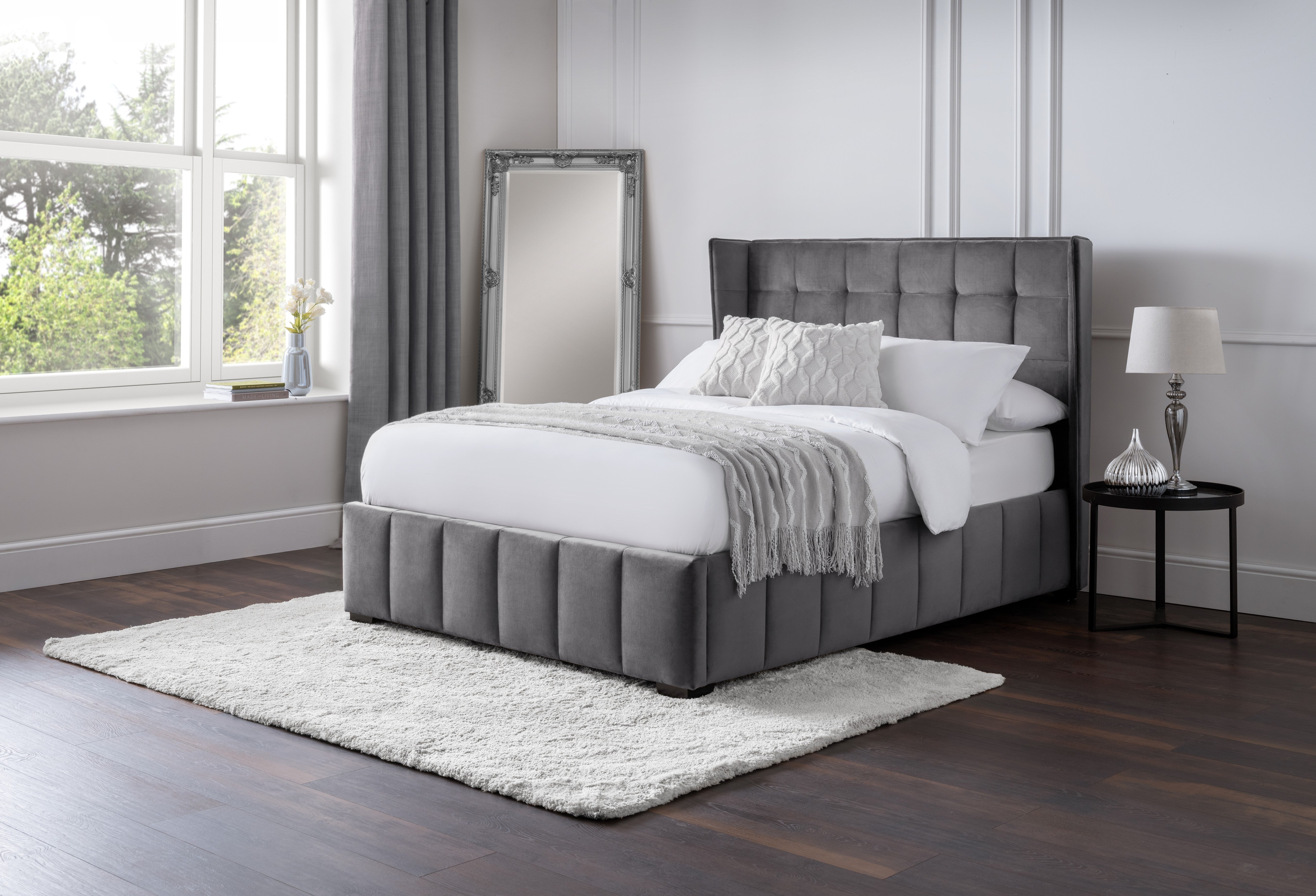 Gray upholstered bed in a bedroom with a window and side table.