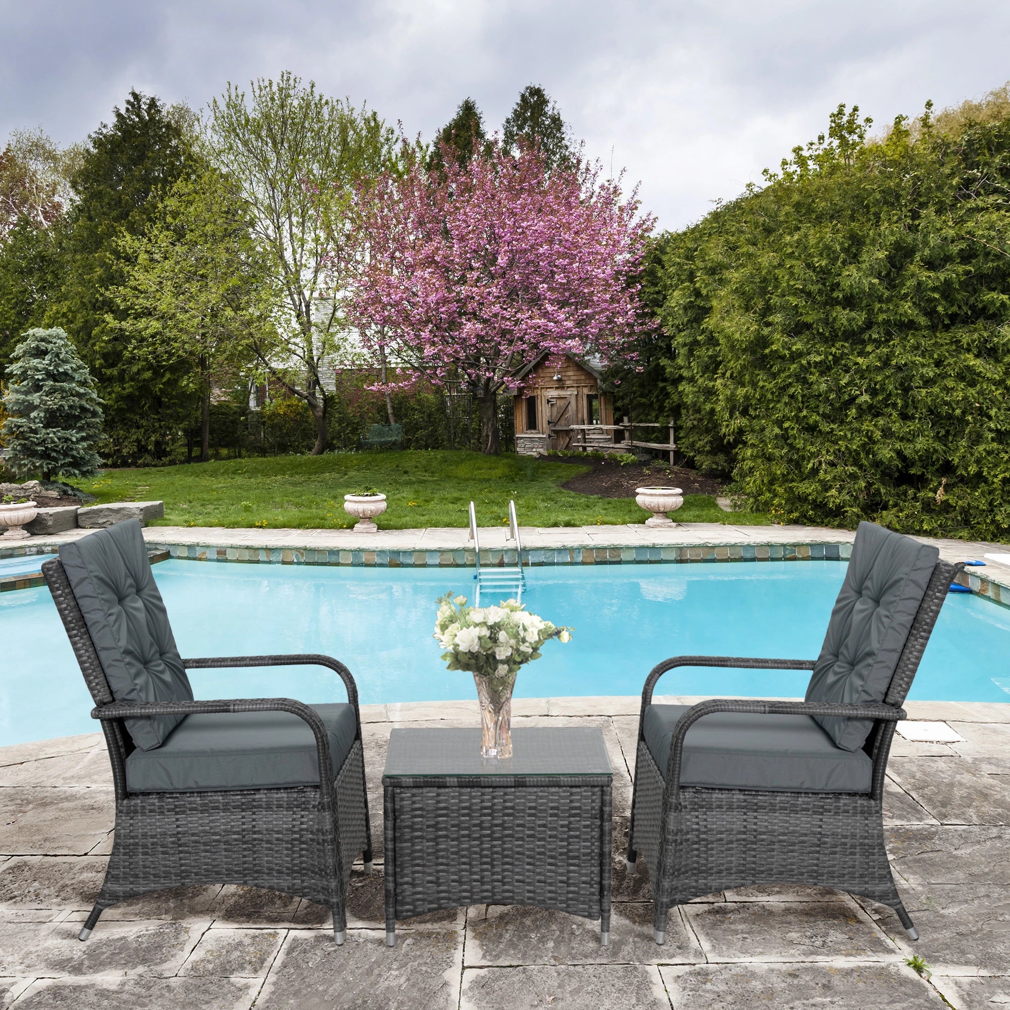 Two wicker chairs with gray cushions and a small table by a poolside with a flowering tree in the background.