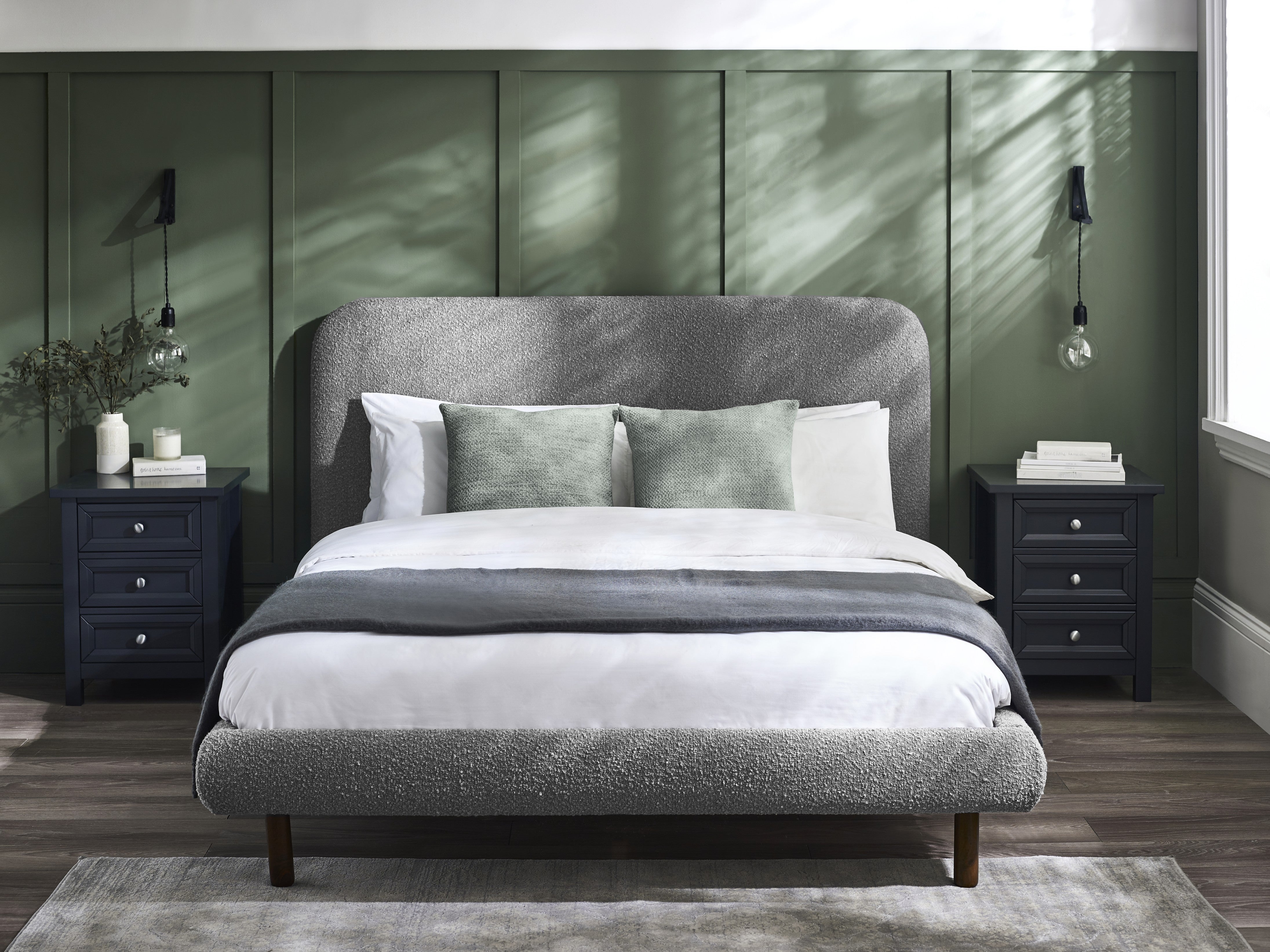 Gray upholstered bed with white bedding in a bedroom setting with green walls and dark nightstands.