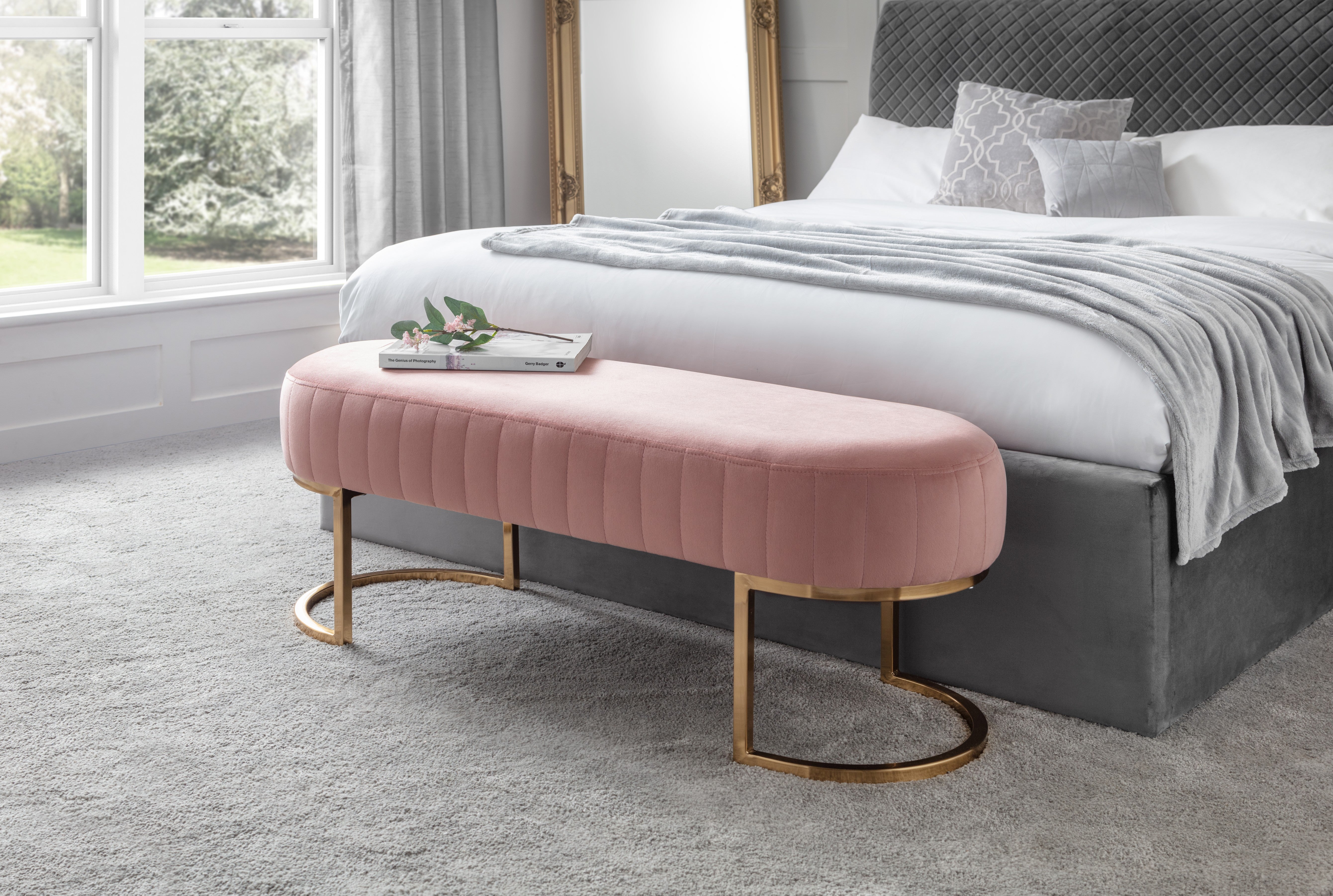 Pink ottoman with gold legs in front of a bed with gray bedding.