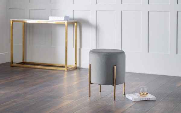 Gray ottoman with gold legs on a wooden floor against a white paneled wall.