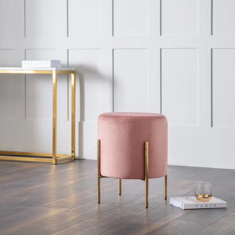 Pink stool with gold legs in a room with a white paneled wall and wooden floor.