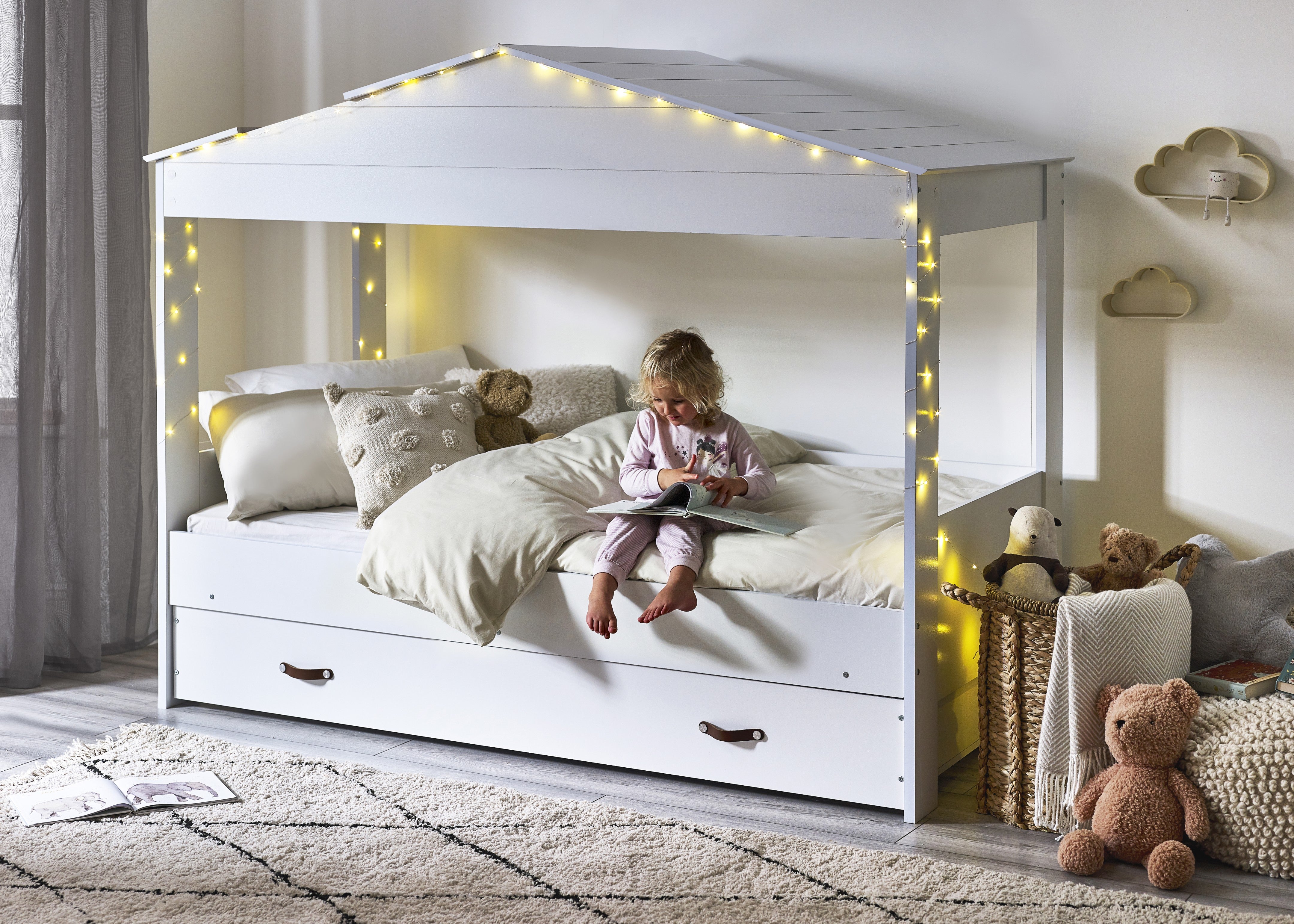 Child reading on a white children's bed with house design, surrounded by toys and soft lighting.