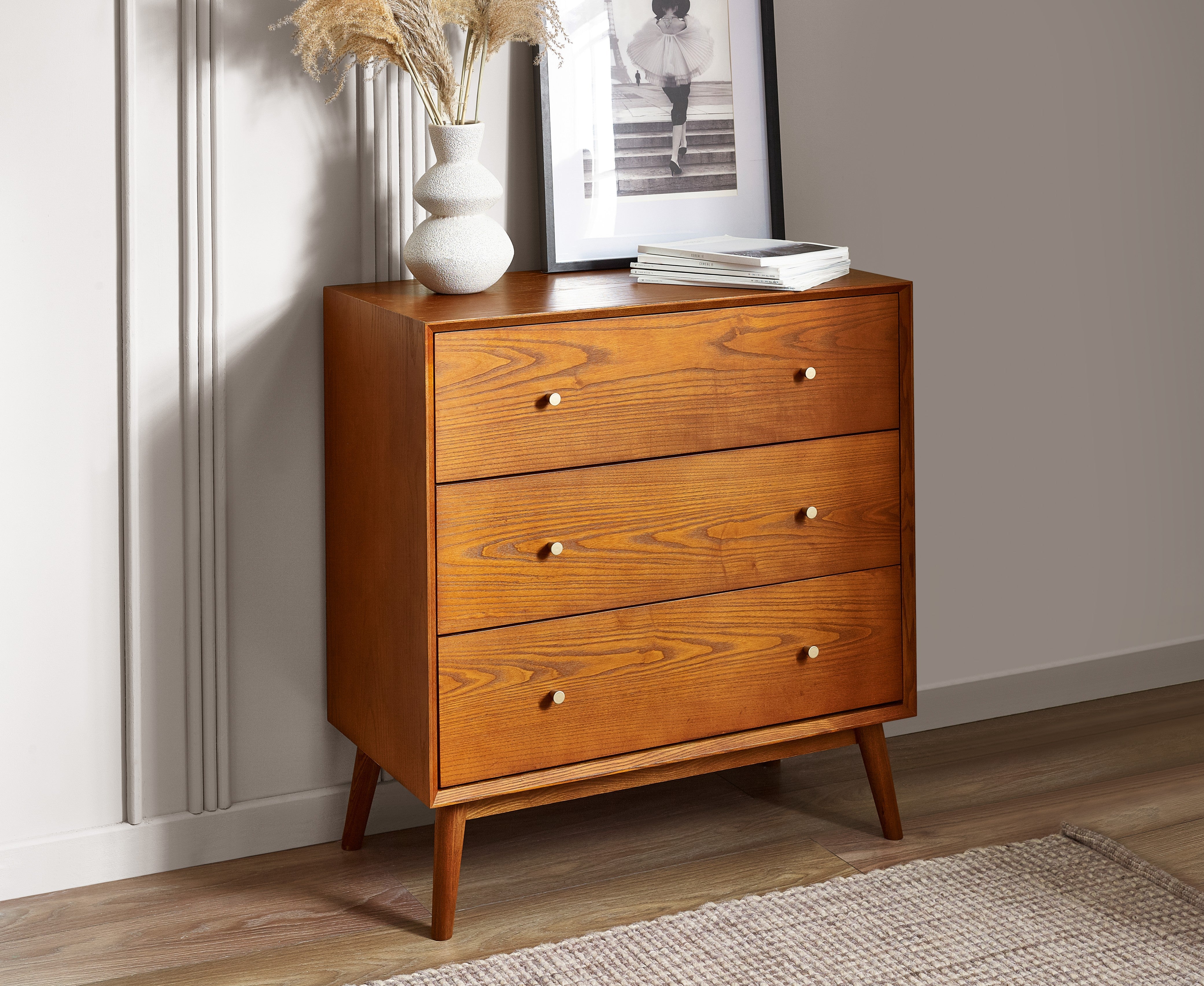 Wooden dresser with three drawers against a wall with a mirror and decor items.