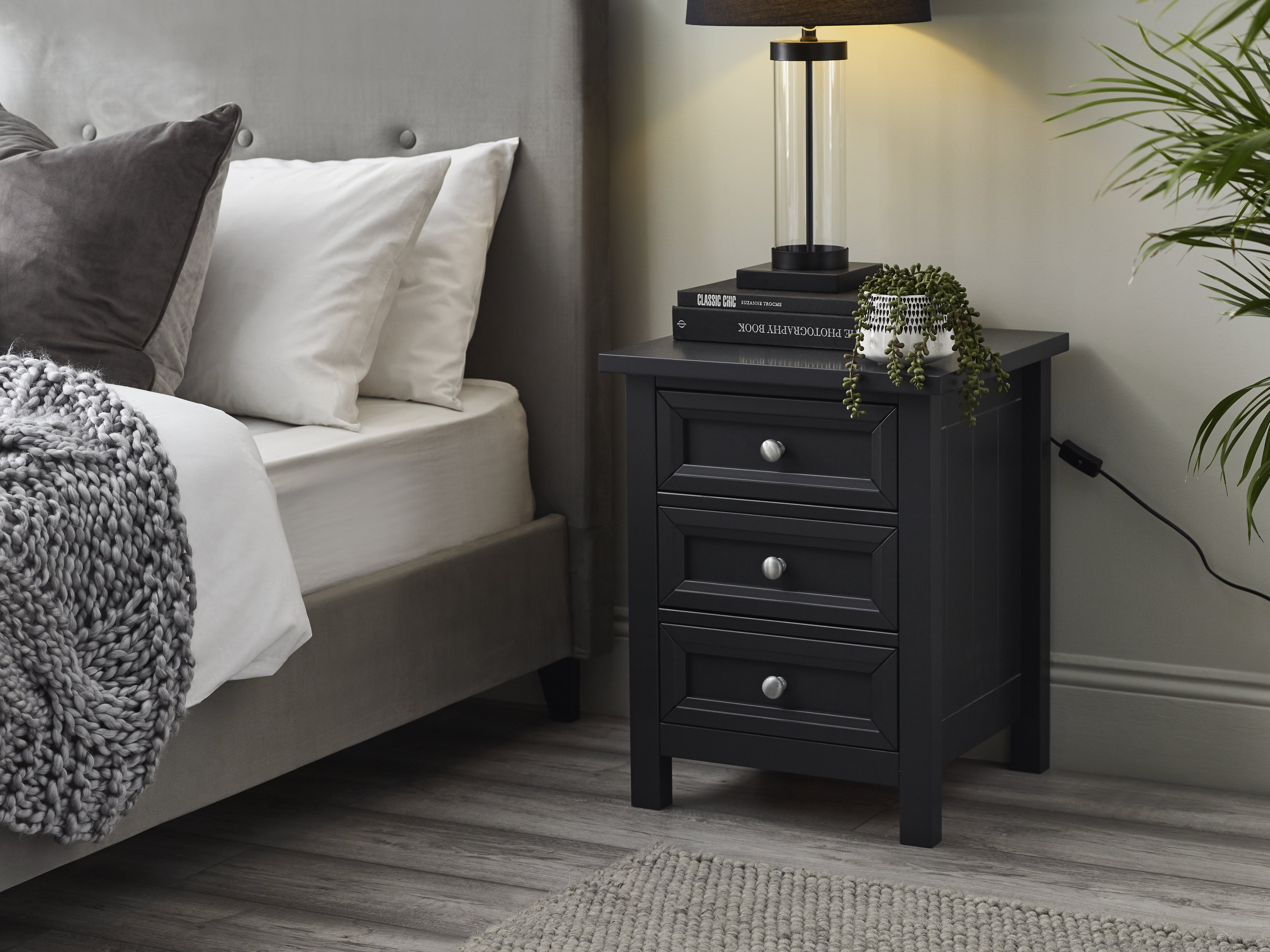 Black nightstand with books and a lamp next to a bed with gray bedding.