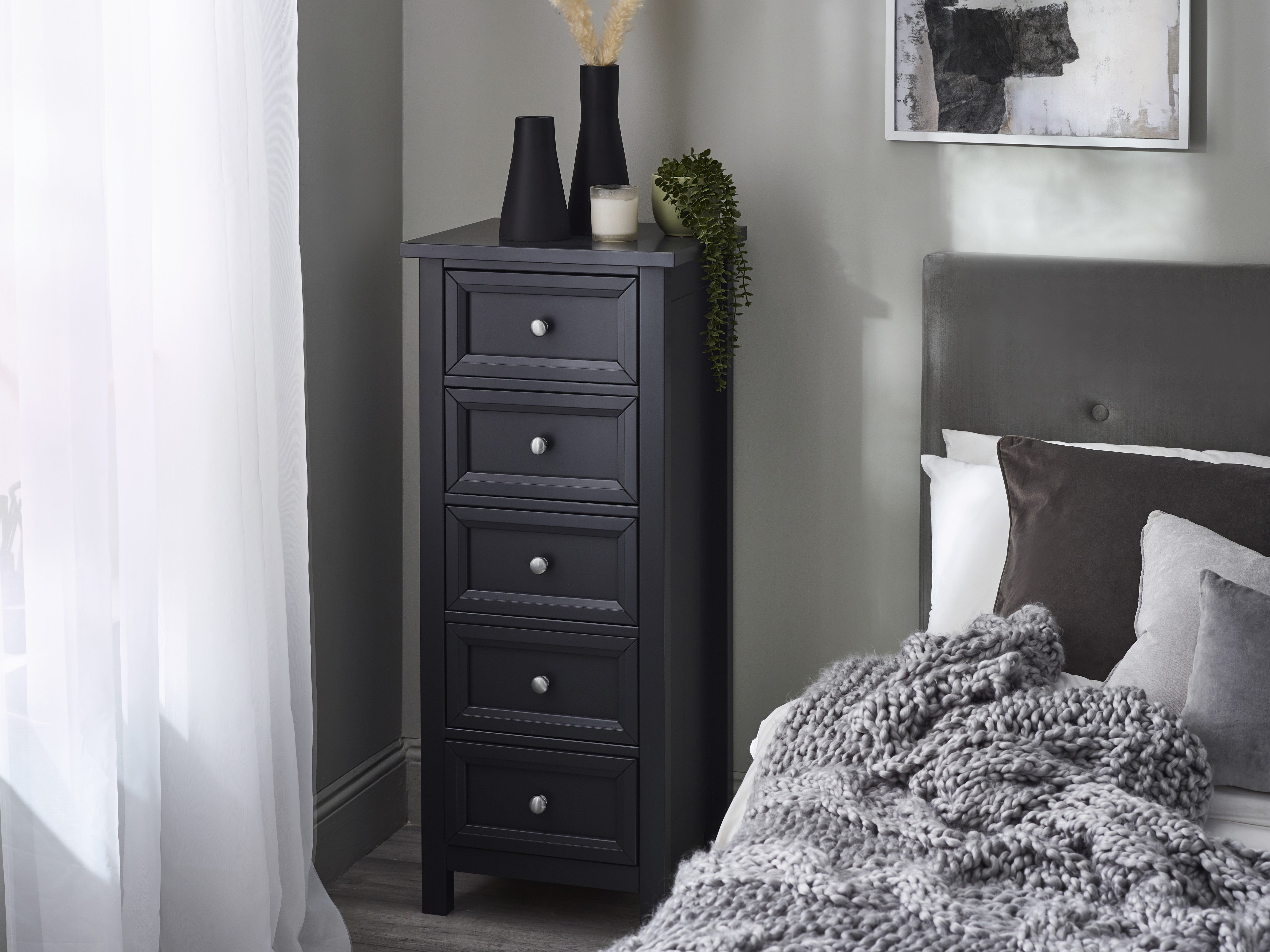 Black tall dresser next to a bed with gray bedding in a bedroom setting.