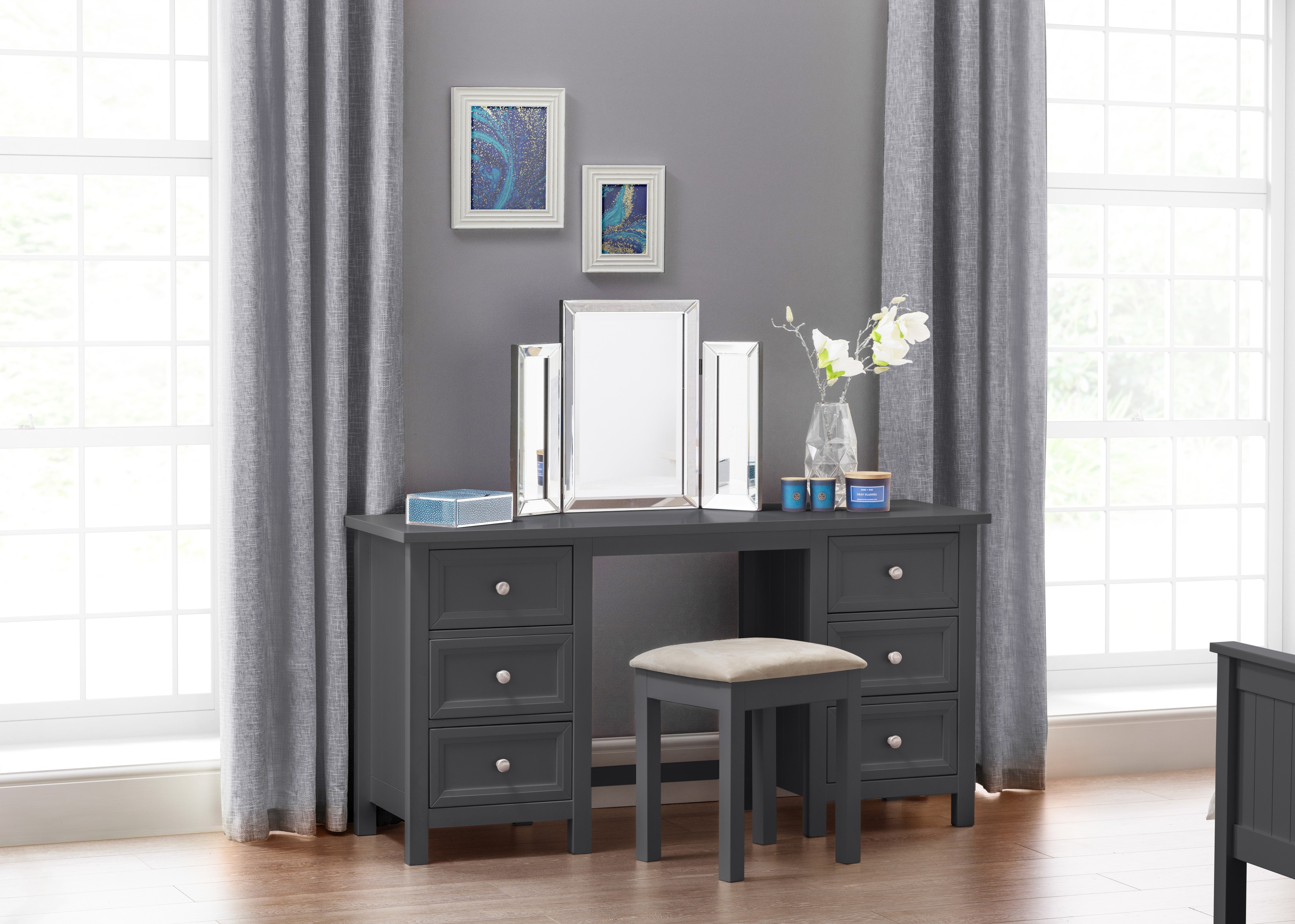 Gray vanity desk with mirror and stool in a room with curtains and framed pictures.