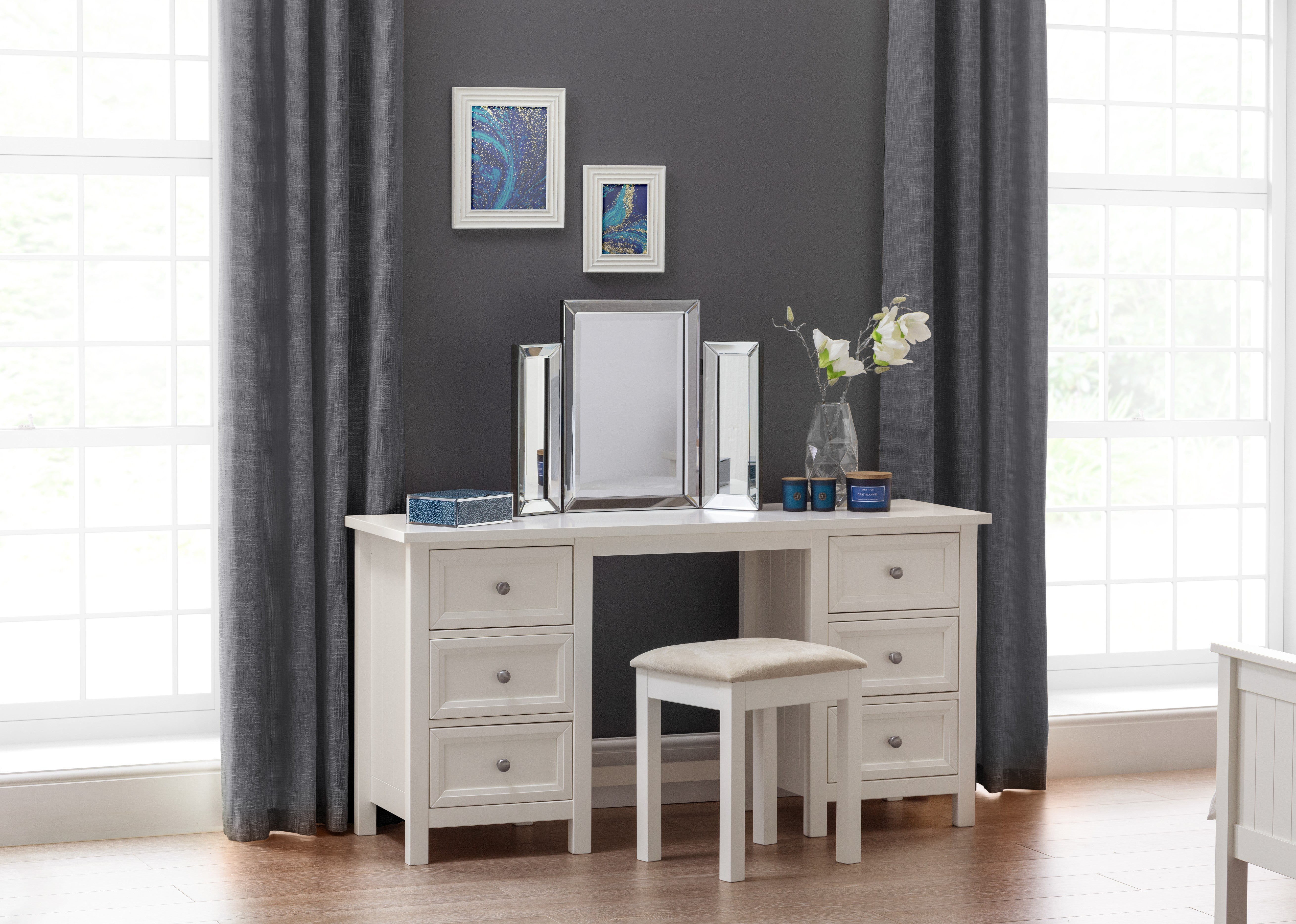 Dressing table with mirror, stool, and decorative items in a room with gray walls and large windows.