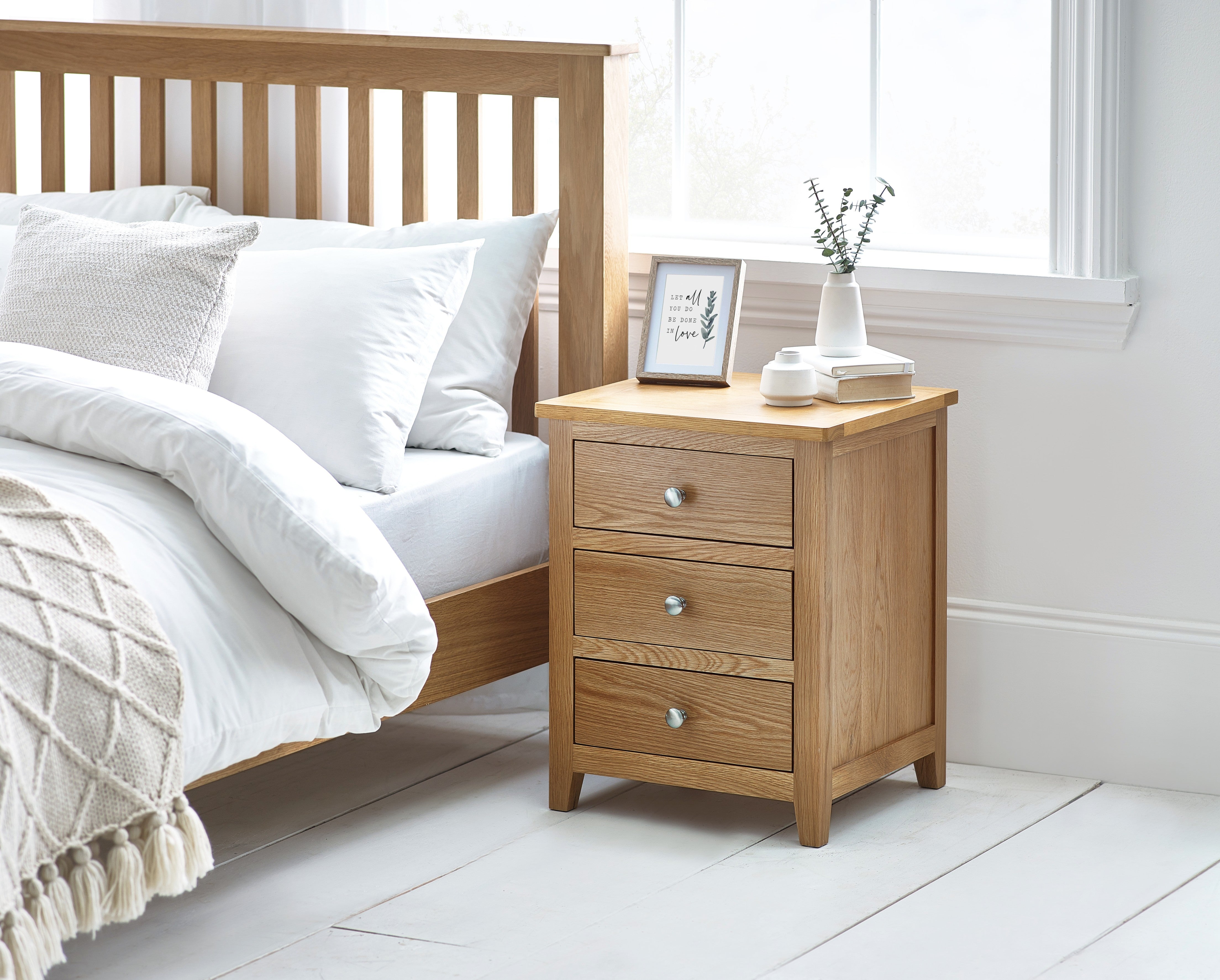 Wooden bedside table with three drawers next to a bed in a bright bedroom.