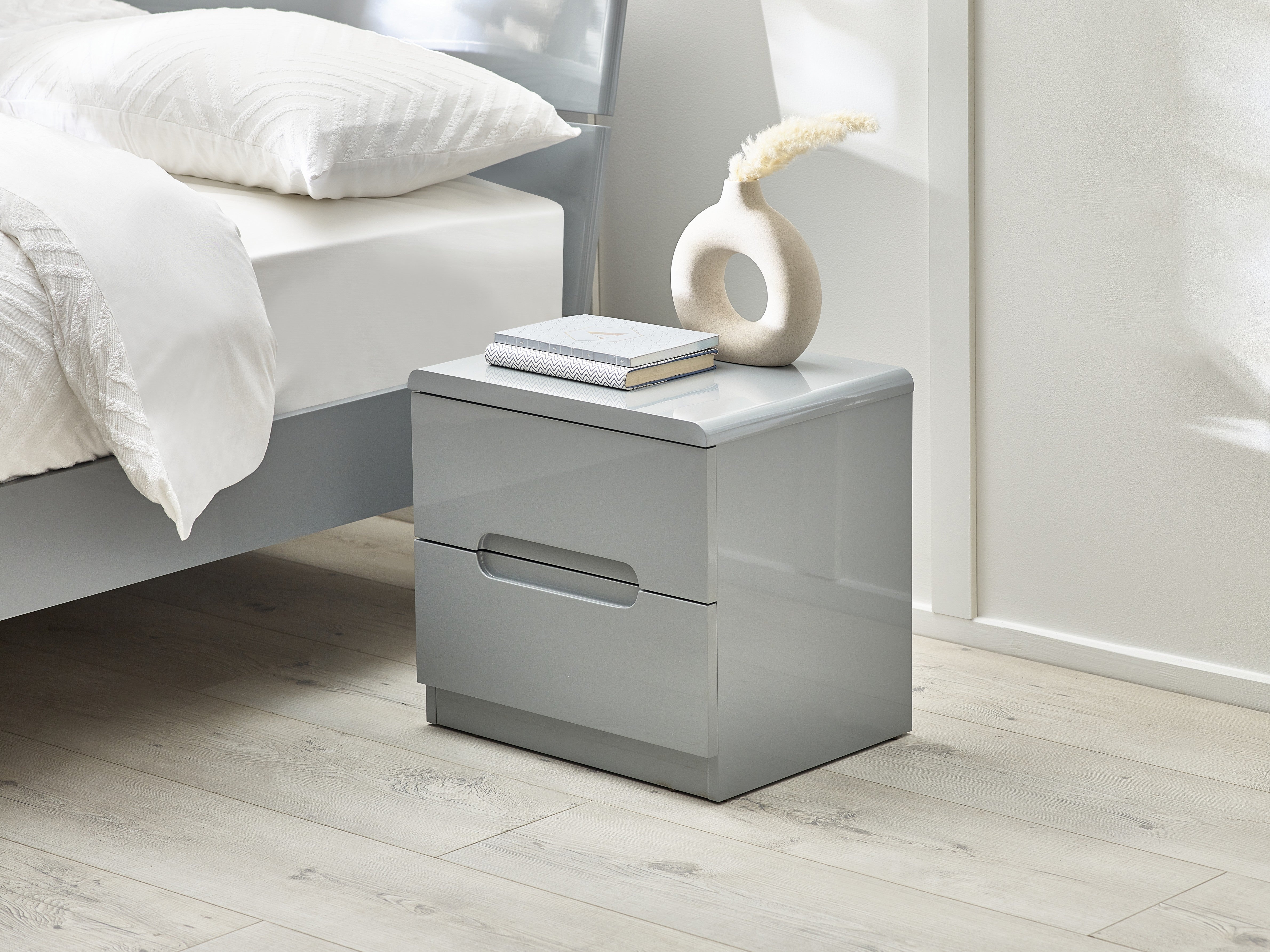 Gray nightstand with a white vase and book next to a bed in a bedroom.