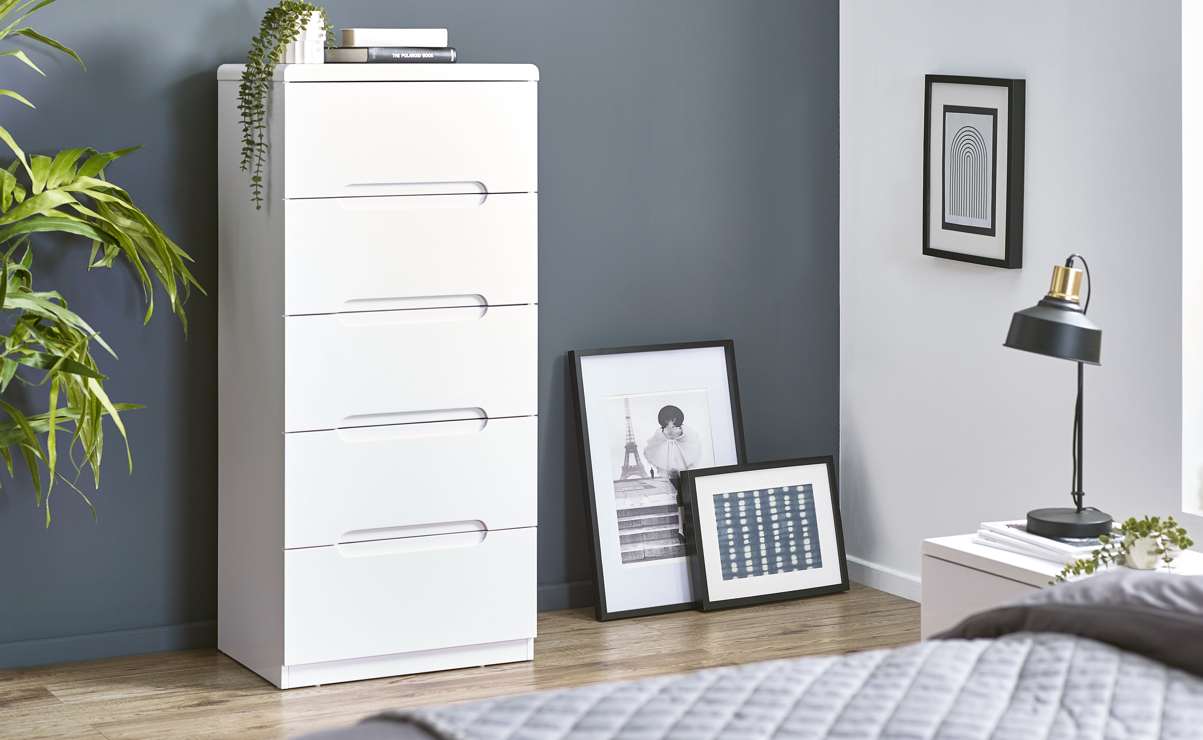 White dresser against a gray wall with framed pictures and a lamp on a nightstand.