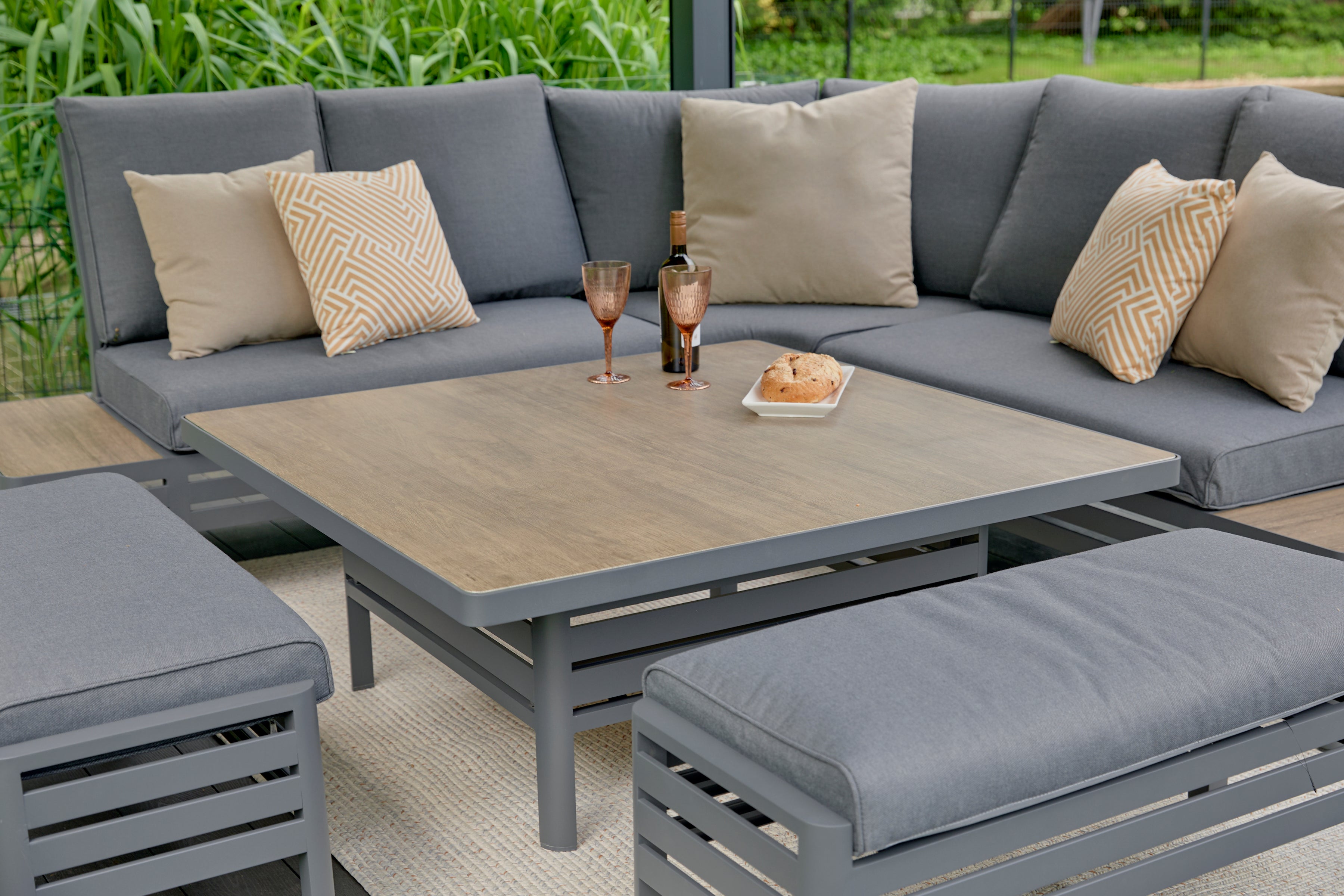 Gray outdoor sectional with cushions and a table, set against a green garden background.