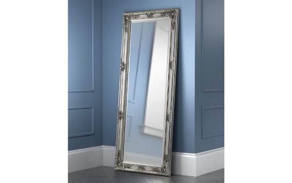 Decorative floor mirror with silver frame leaning against a blue wall.