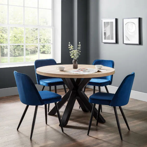Round wooden dining table with four blue chairs in a room with large windows.