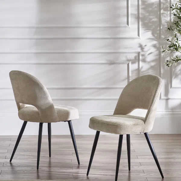 Two beige chairs with black legs on a wooden floor against a white paneled wall.