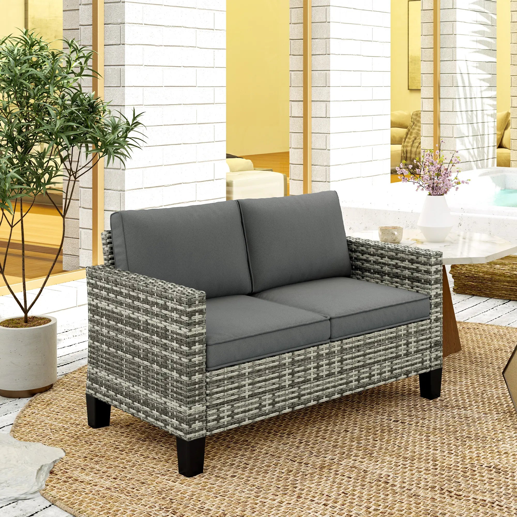 Gray wicker loveseat with dark cushions on a patio with plants and furniture.