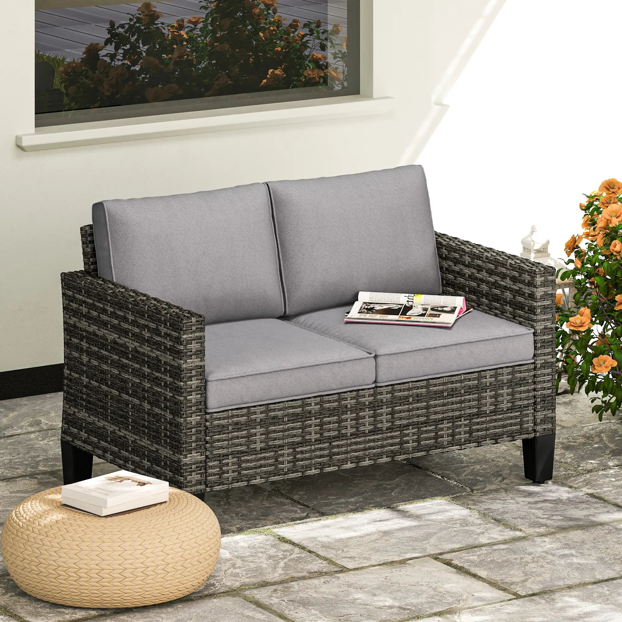 Wicker outdoor loveseat with gray cushions on a patio, next to a small table and flowers.