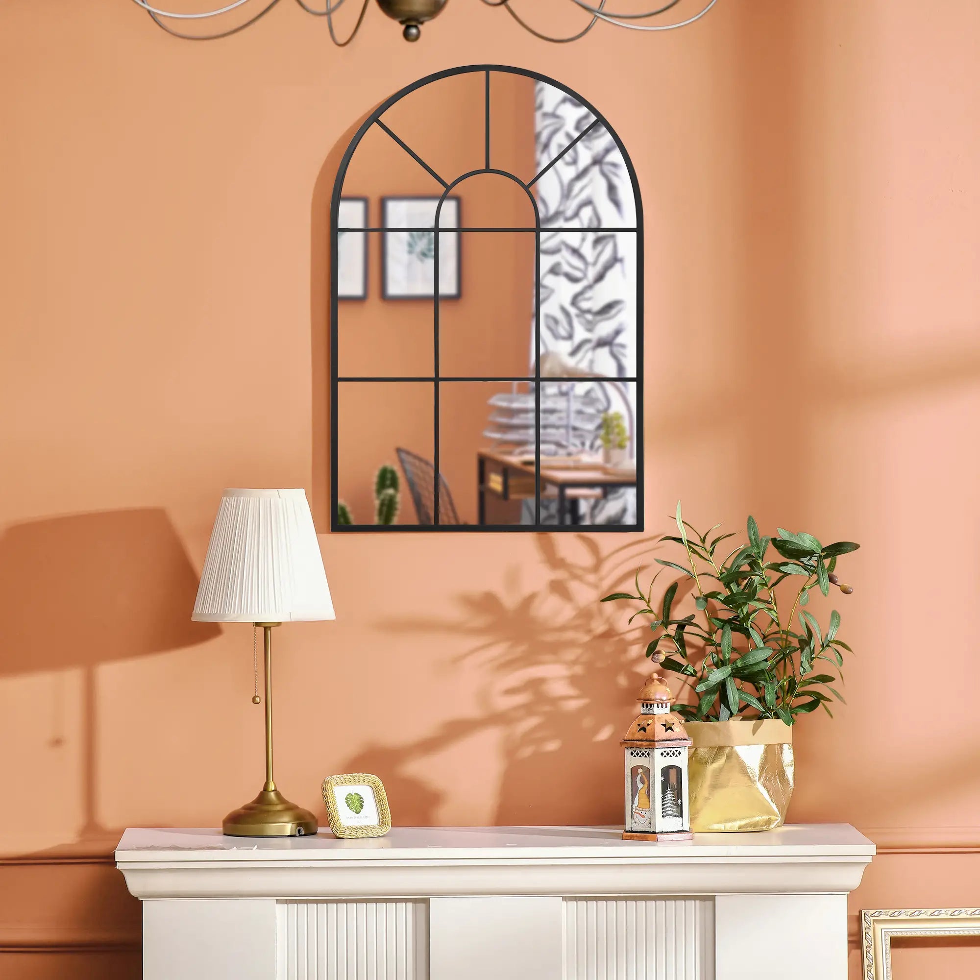 Decorative mirror on a peach wall with a lamp, plant, and lantern on a white console table.